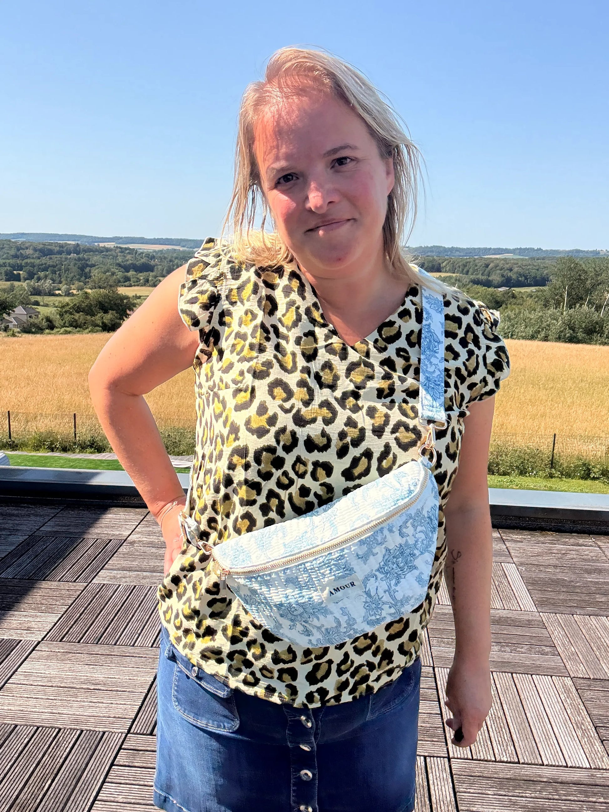 Sac banane Toile de Jouy – Imprimé bleu turquoise portée en bandoulière par une femme sur une terrasse, paysage champêtre en arrière-plan.