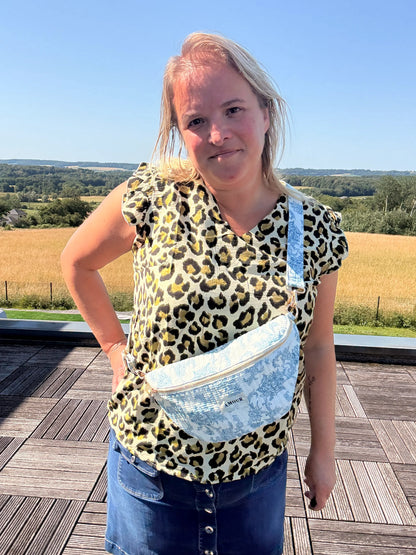 Sac banane Toile de Jouy – Imprimé bleu turquoise portée en bandoulière par une femme sur une terrasse, paysage champêtre en arrière-plan.