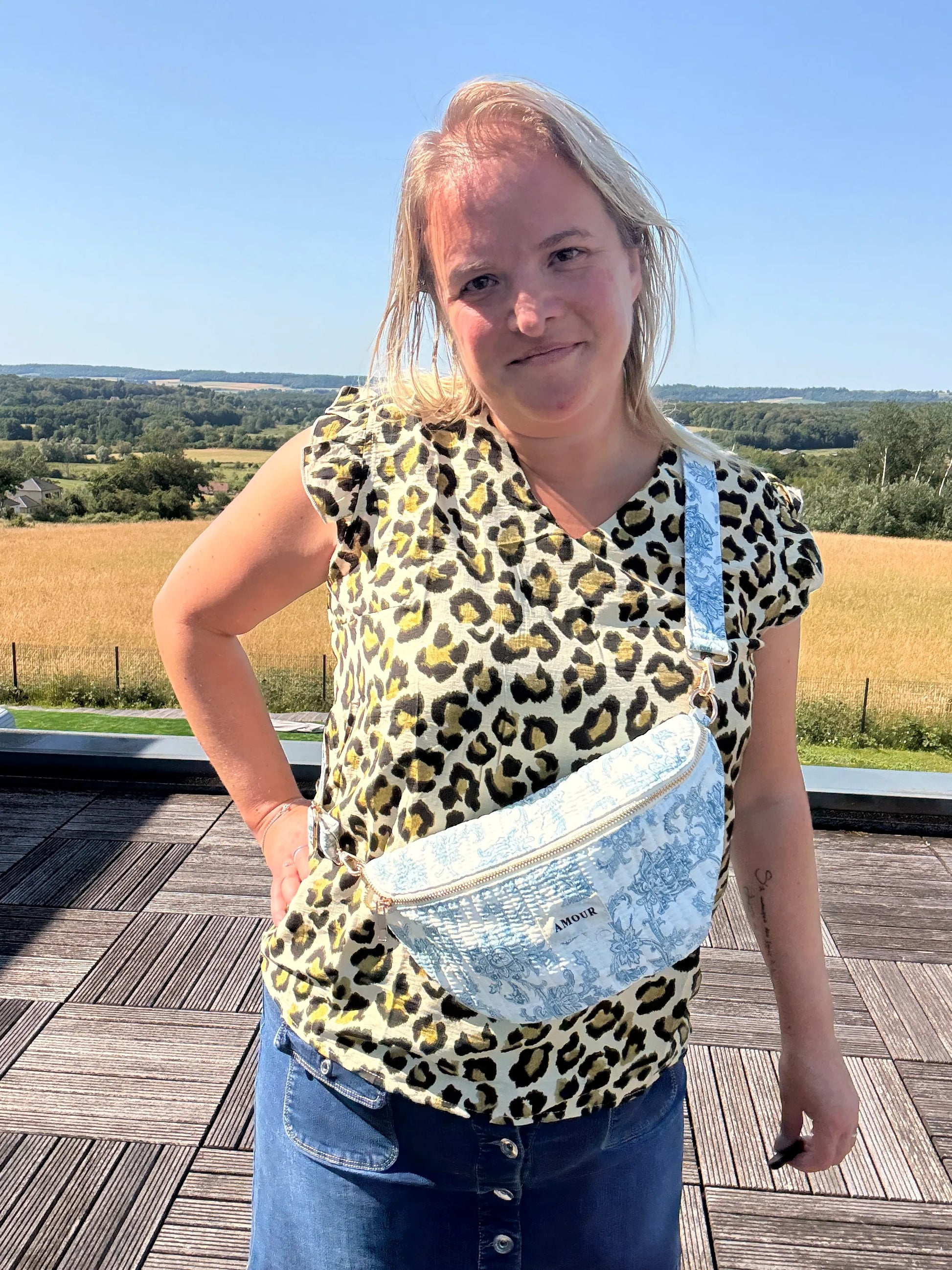 Personne portant un sac banane Toile de Jouy imprimé bleu turquoise, en outdoors sur fond champ, chemise à motif léopard et jean.