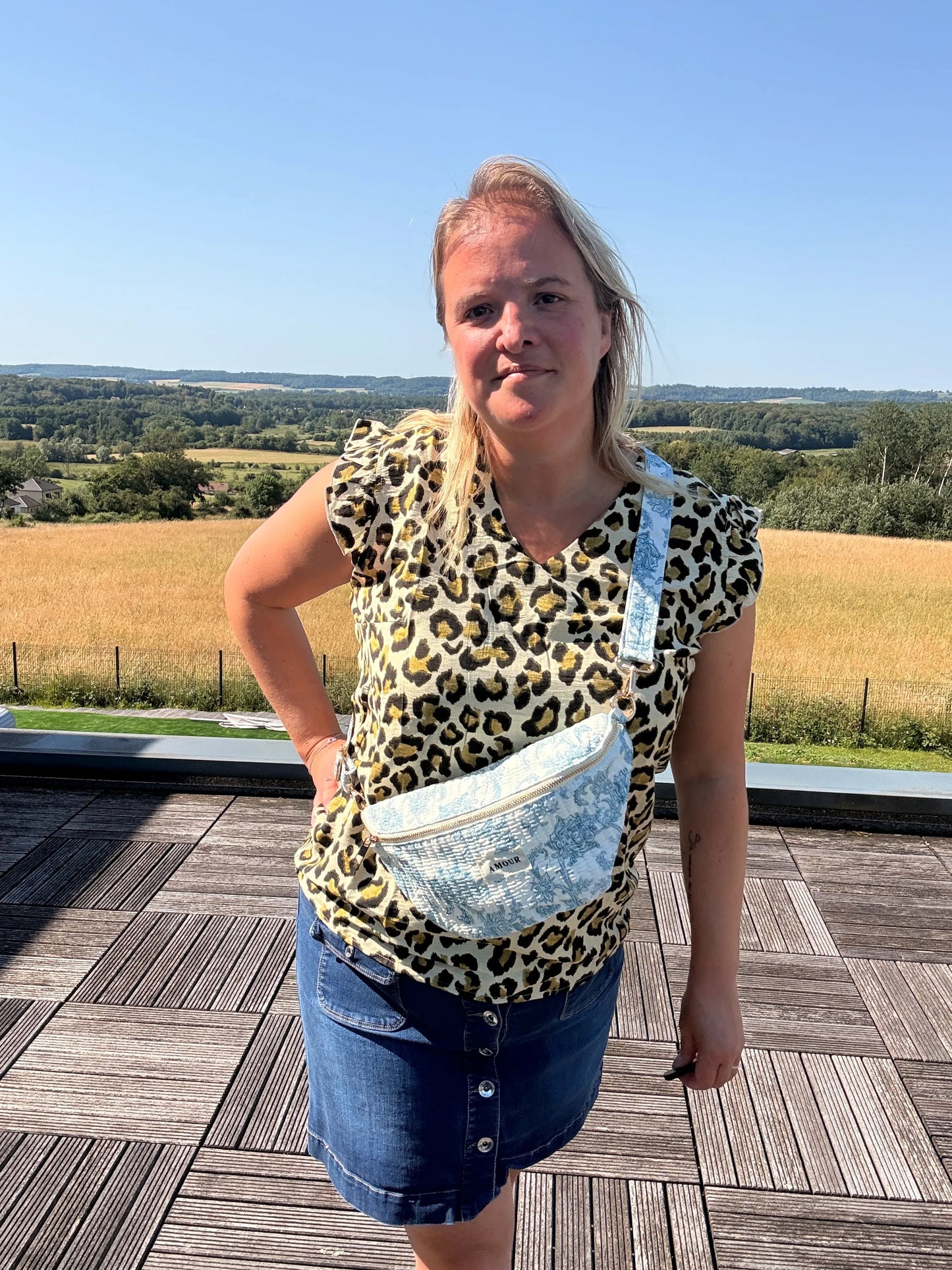 Homme/femme portant un sac banane Toile de Jouy bleu turquoise, posé sur la poitrine, silhouette debout sur une terrasse en plein air avec champ et ciel bleu.