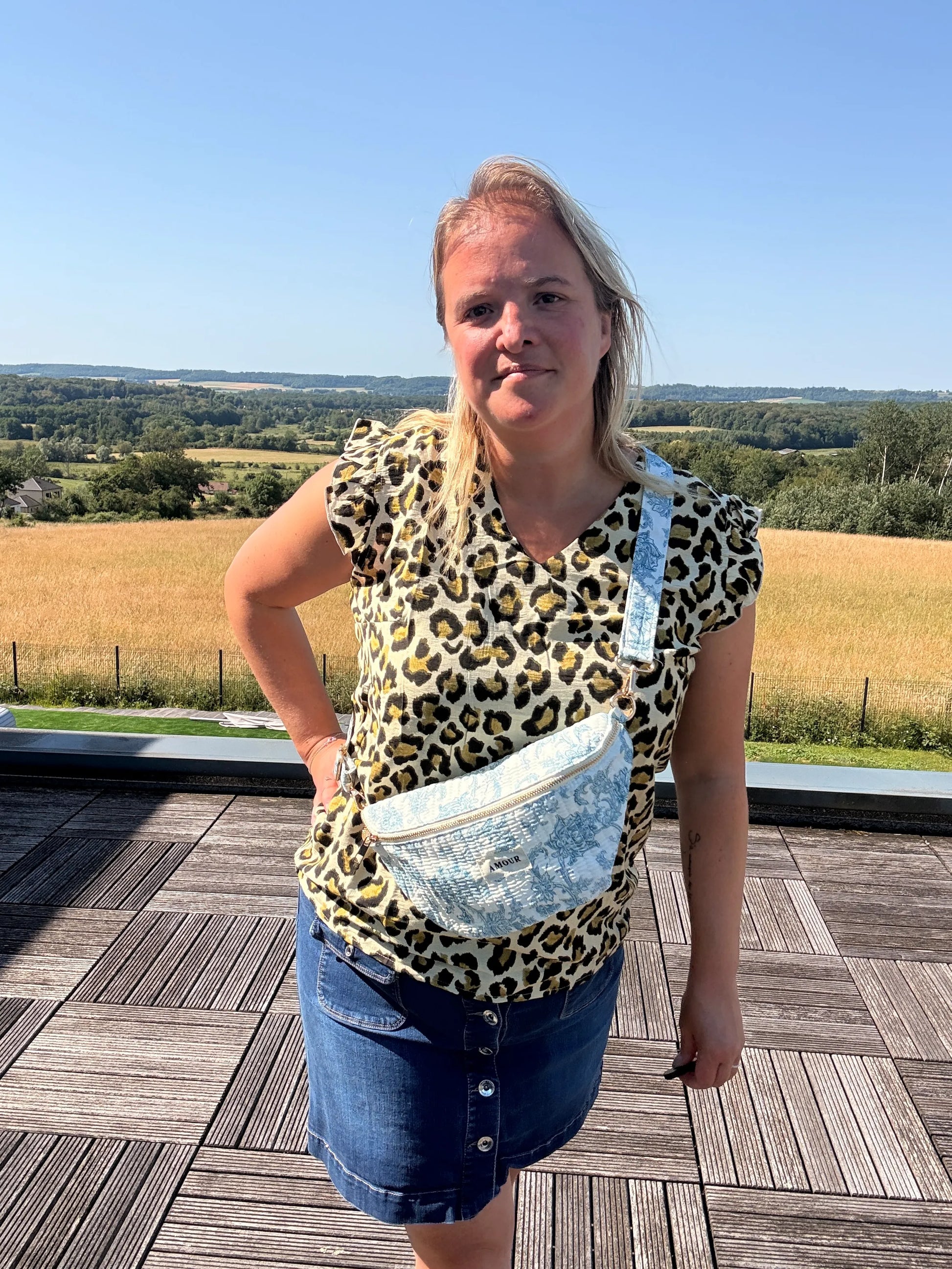 Homme/femme portant un sac banane Toile de Jouy bleu turquoise, posé sur la poitrine, silhouette debout sur une terrasse en plein air avec champ et ciel bleu.