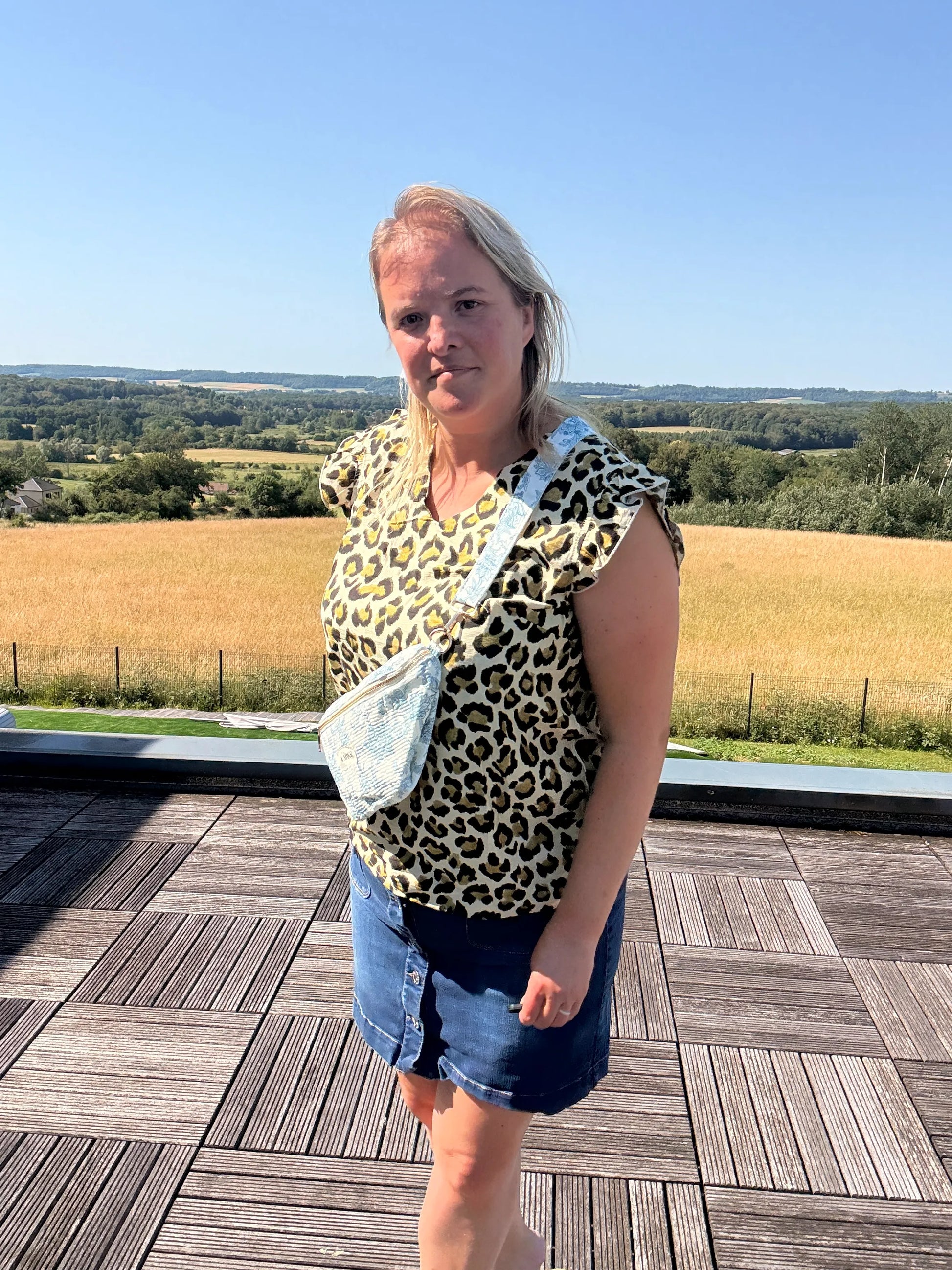 Sac Banane Toile de Jouy – Imprimé Bleu Turquoise: femme portant top léopard, short en jean, sac banane bleu turquoise crossbody sur terrasse avec champ et ciel bleu.