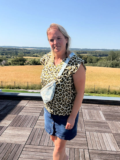 Sac Banane Toile de Jouy – Imprimé Bleu Turquoise: femme portant top léopard, short en jean, sac banane bleu turquoise crossbody sur terrasse avec champ et ciel bleu.
