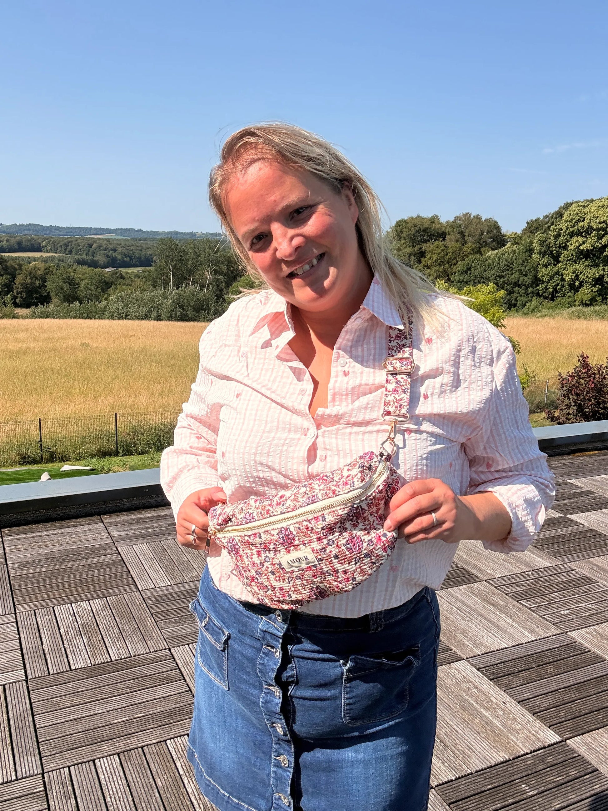 Sac Banane Liberty Rose – Imprimé Fleuri Femme: femme souriante porte une banane en tissu imprimé fleurs rose et blanc, sur fond paysage champêtre.