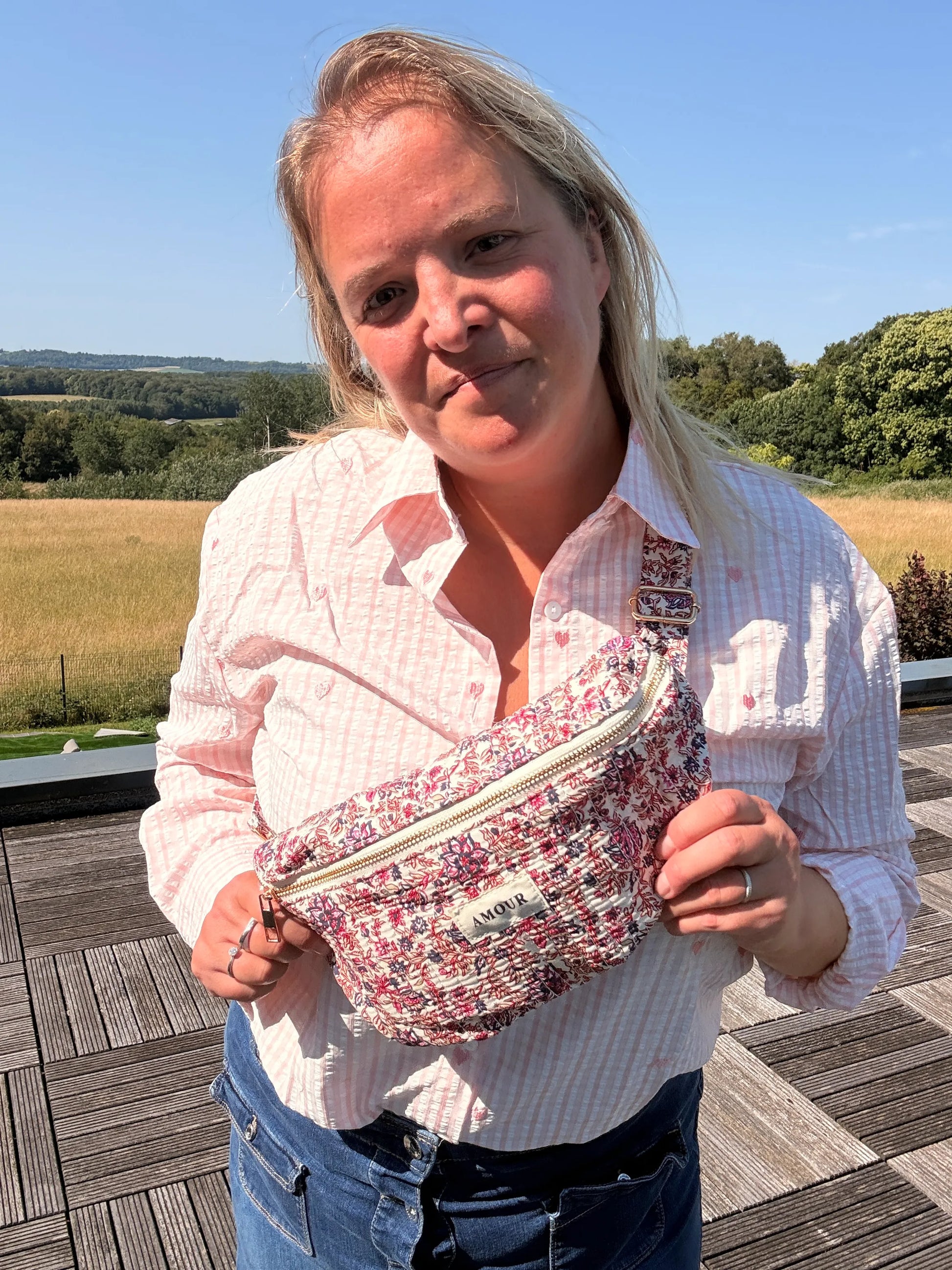 Femme souriante portant un sac banane Liberty rose à imprimé fleuri, posé sur le torse en plein air.