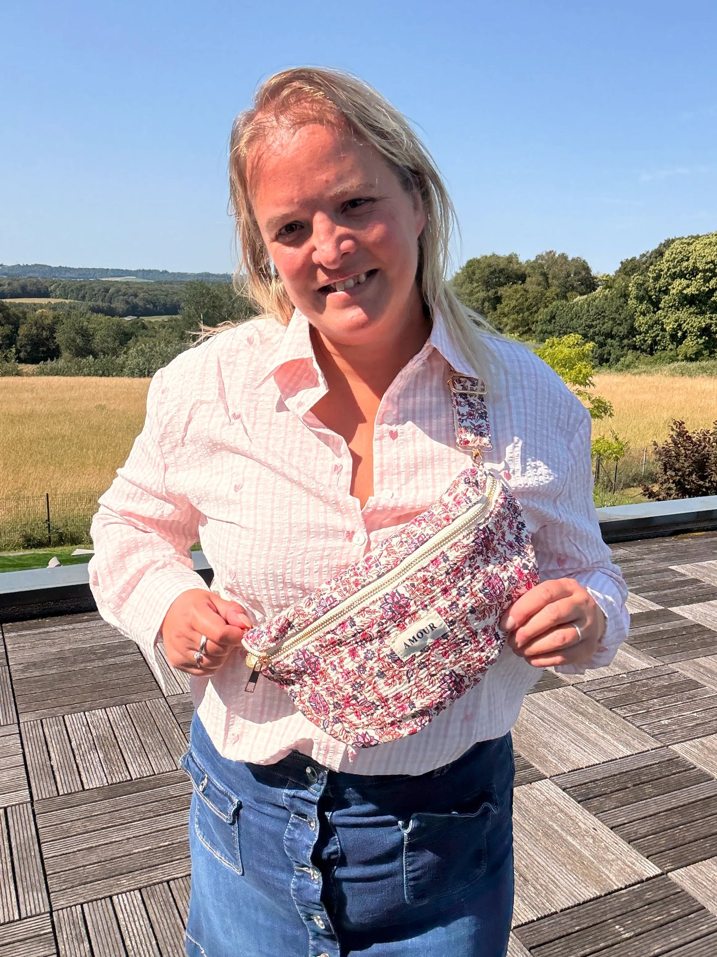 Femme portant un sac banane Liberty rose à imprimé fleuri sur une chemise rose clair, en extérieur sunny sur fond de paysage champêtre.