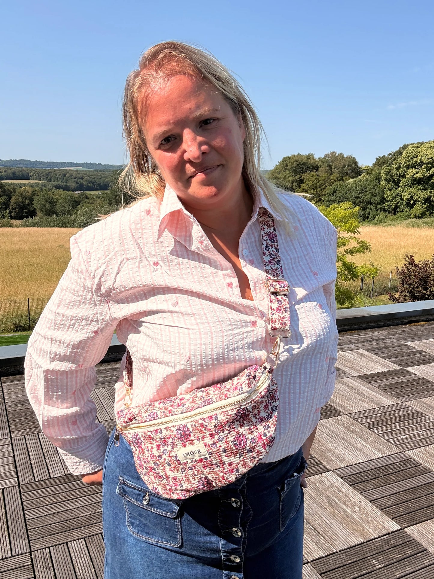 Personne portant un sac banane Liberty rose imprimé fleuri, tenue chemise rose et jean, debout en extérieur sur une terrasse.