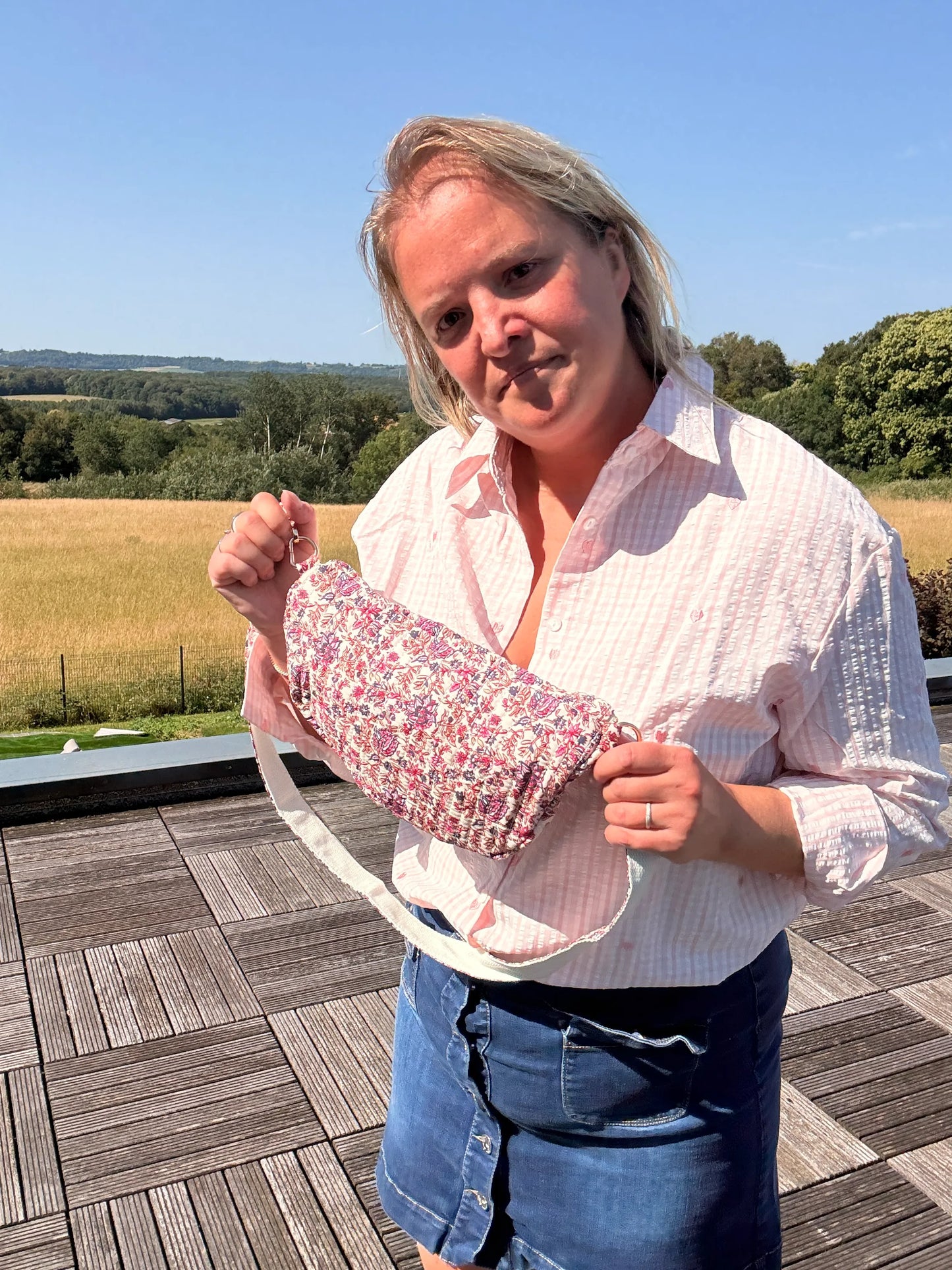 Femme tenant un sac banane Liberty Rose imprimé fleuri, près d’un espace extérieur, sur fond de paysage champêtre.