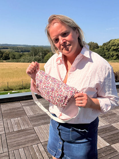 Femme tenant un sac banane Liberty Rose imprimé fleuri, près d’un espace extérieur, sur fond de paysage champêtre.