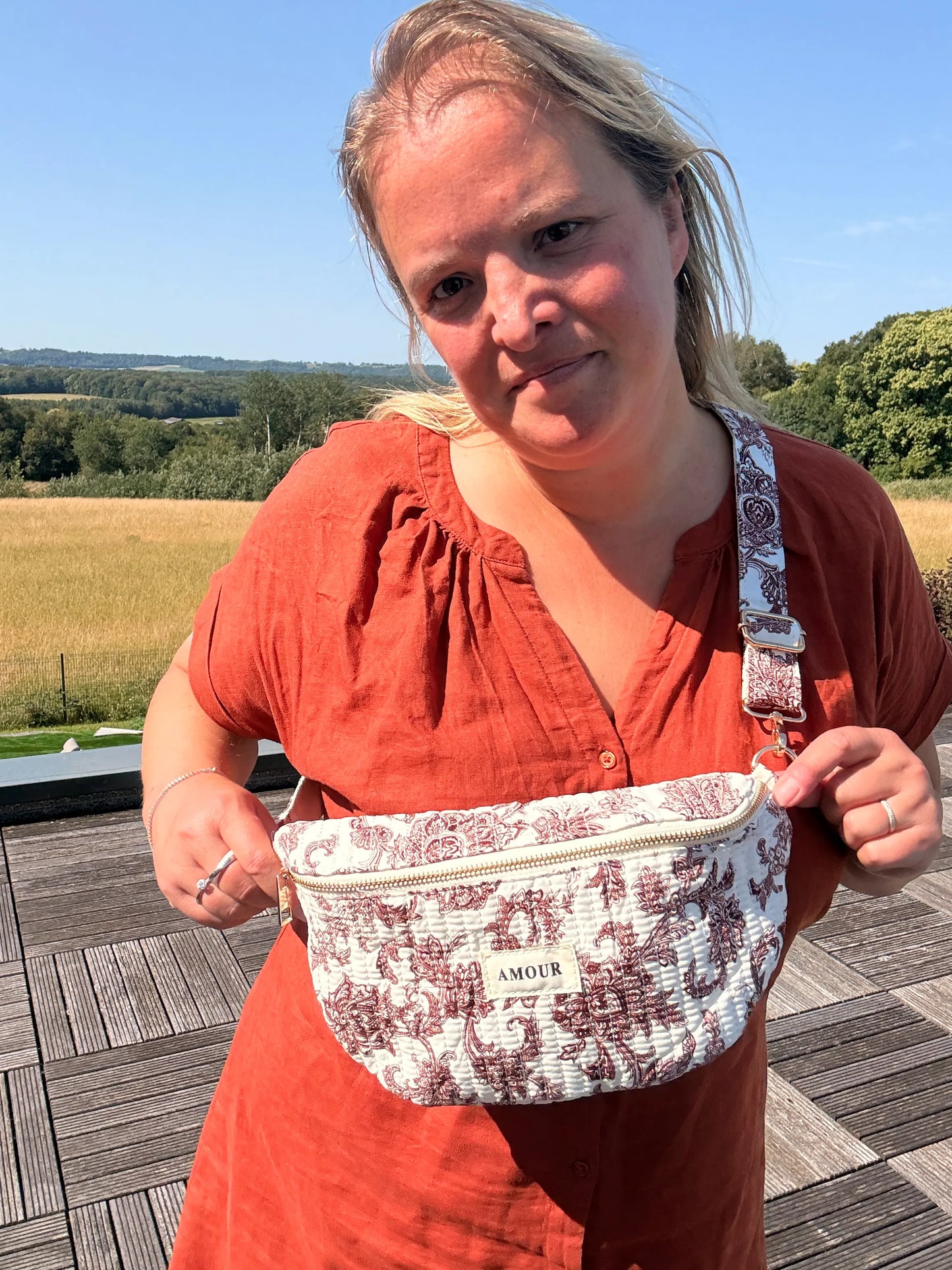 Sac banane en toile de Jouy Bordeaux imprimé Amour posé sur l’épaule d’une femme, devant un paysage champêtre.