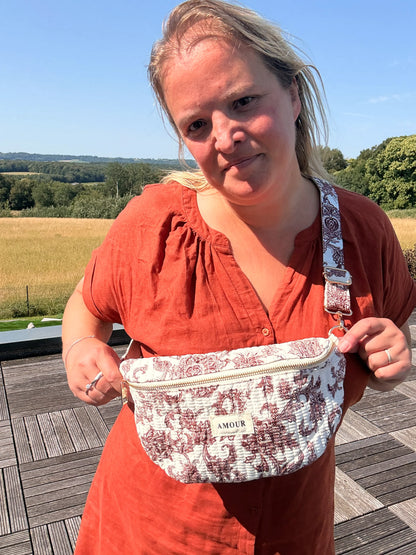 Sac banane en toile de Jouy Bordeaux imprimé Amour posé sur l’épaule d’une femme, devant un paysage champêtre.