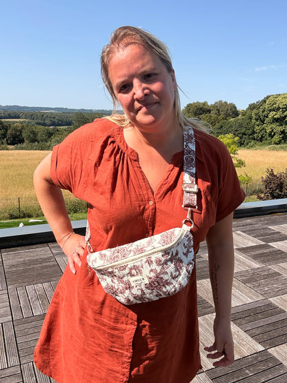 Sac banane Toile de Jouy Bordeaux imprimé Amour porté en bandoulière sur une femme en robe orange sur une terrasse rurale.