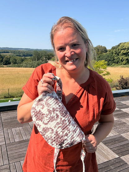 Femme tenant un sac banane en Toile de Jouy Bordeaux imprimé Amour, sourire sur fond de paysage champêtre.