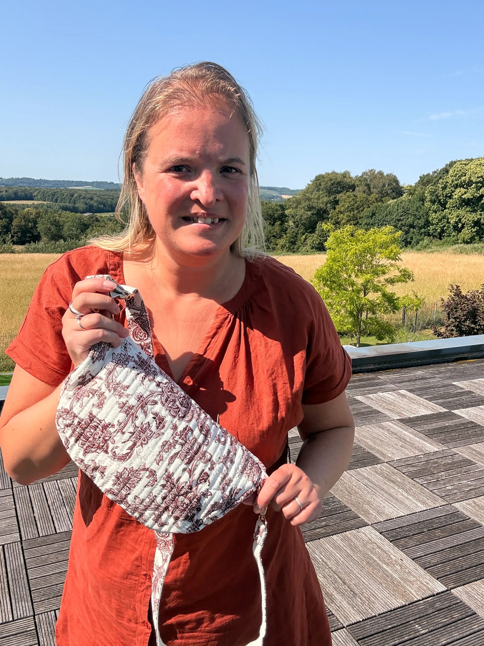 Femme souriante portant un sac banane imprimé Toile de Jouy Bordeaux, tenue orange, en extérieur sur une terrasse avec champ et arbres au loin.