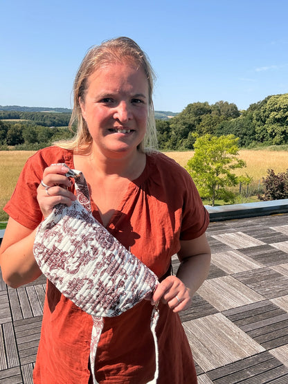 Femme souriante portant un sac banane imprimé Toile de Jouy Bordeaux, tenue orange, en extérieur sur une terrasse avec champ et arbres au loin.