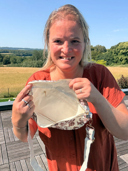 Sac banane en toile de Jouy Bordeaux, montrant l’intérieur clair et une fermeture éclair; femme tenant le sac ouverte en extérieur, paysage champêtre en arrière-plan.
