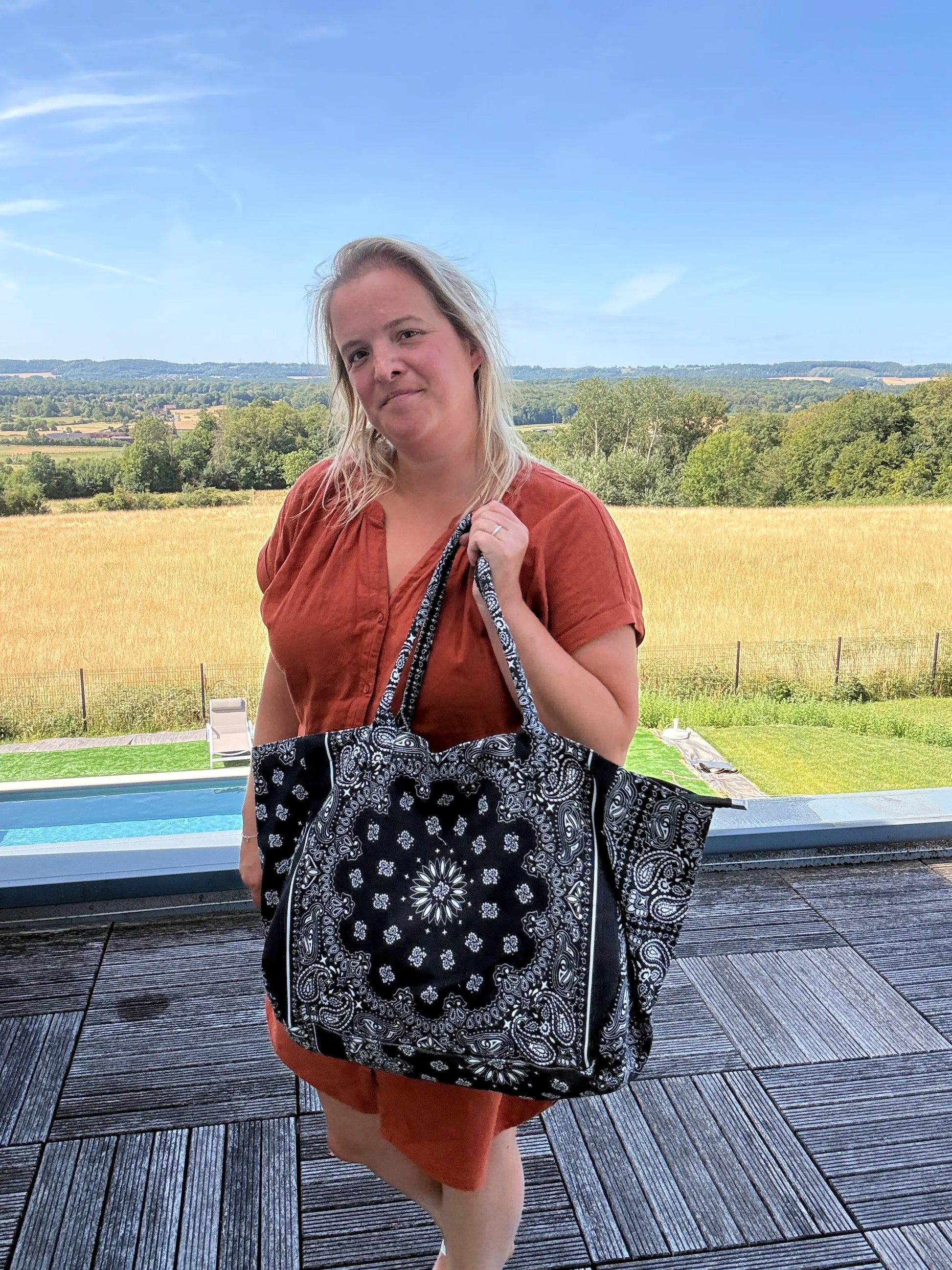 Sac polochon noir à motifs paisley porté sur l’épaule par une femme, en robe rouge, près d’une terrasse avec paysage rural en arrière-plan.