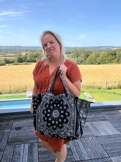 Sac polochon noir à motifs paisley porté sur l’épaule par une femme, en robe rouge, près d’une terrasse avec paysage rural en arrière-plan.