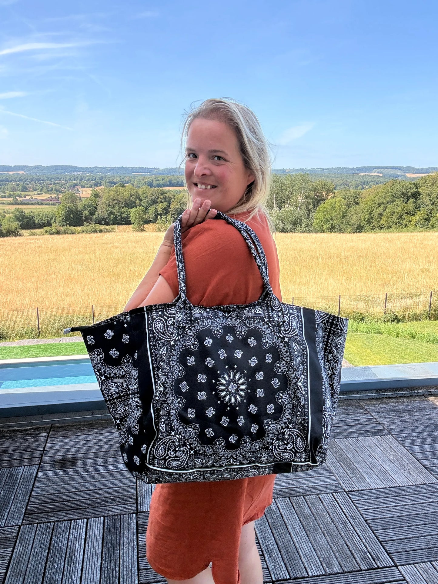 Sac polochon épaules noir imprimé paisley porté sur l’épaule d’une femme en robe orange, avec paysage champêtre en arrière-plan.