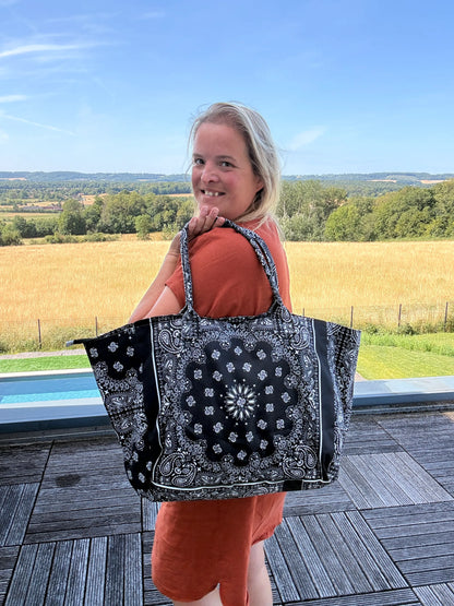 Sac polochon épaules noir imprimé paisley porté sur l’épaule d’une femme en robe orange, avec paysage champêtre en arrière-plan.