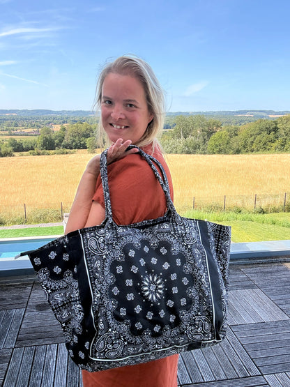 Sac polochon épaule en coton noir à motif paisley porté sur l’épaule, femme souriante en robe orange en arrière-plan campagne. Grand format zippé.