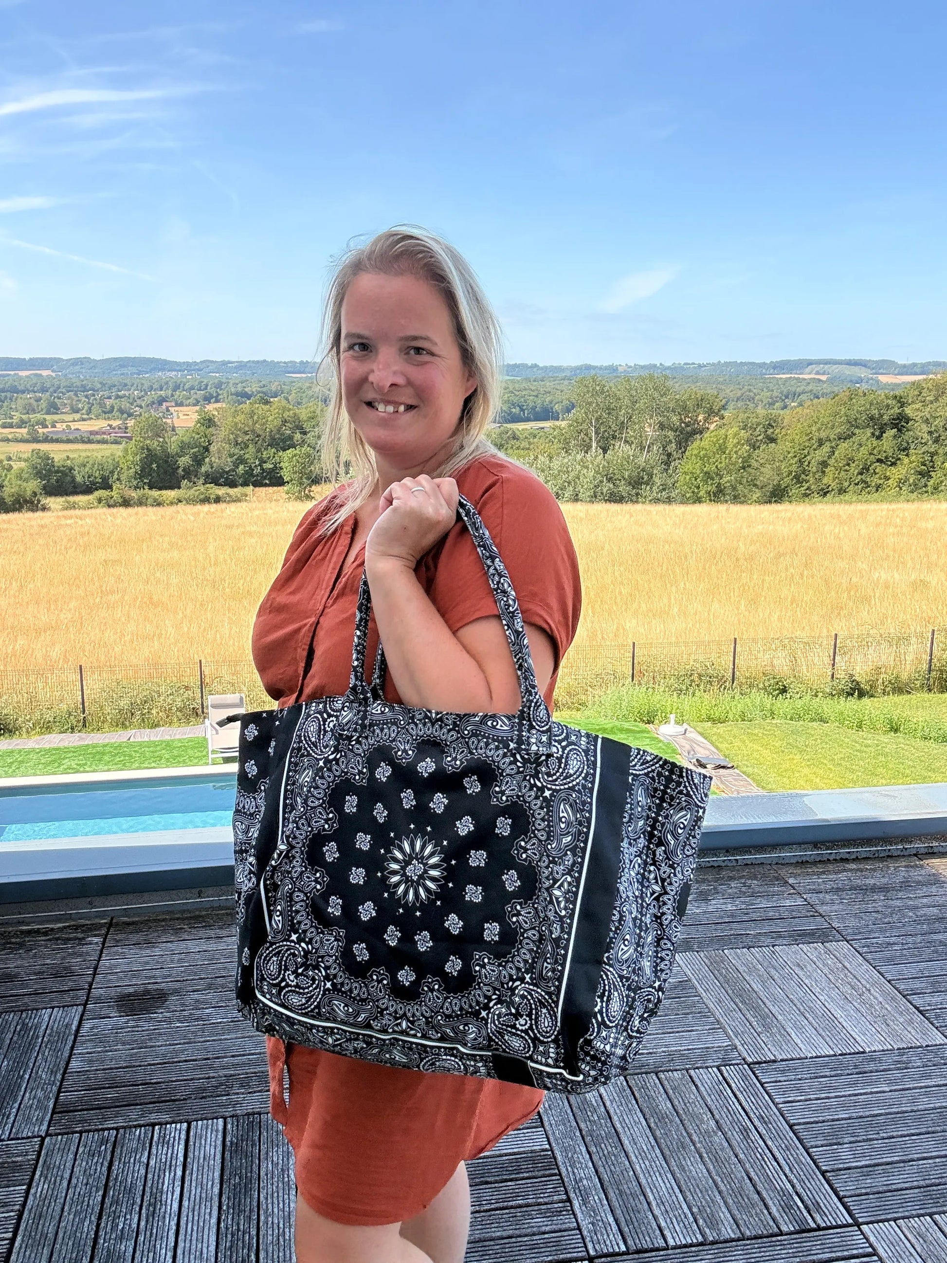 Sac Polochon Épaule Coton Paisley Noir – Grand Format Zippé : une femme souriante porte un grand sac noir à motifs bandana sur fond paisley, en extérieur.