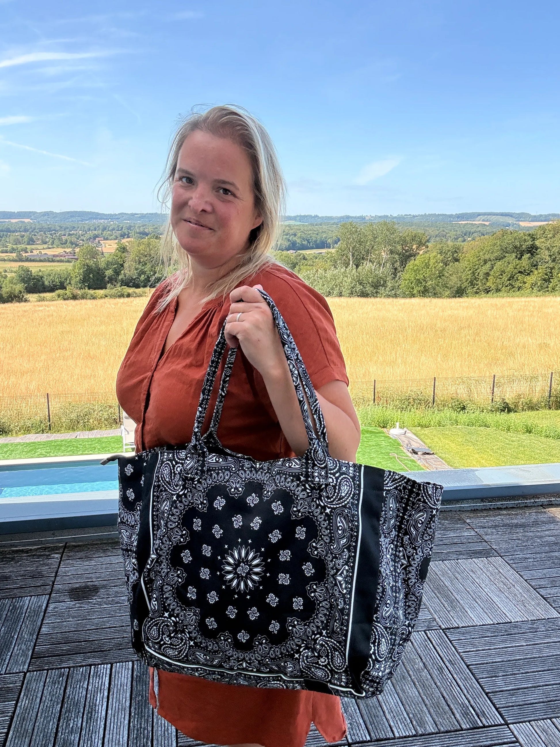 Homme/ femme portant un grand sac polochon noir à motif bandana Paisley, en extérieur près d’une terrasse et d’un paysage champêtre. Nom du produit: Sac Polochon Épaule Coton Paisley Noir – Grand Format Zippé.