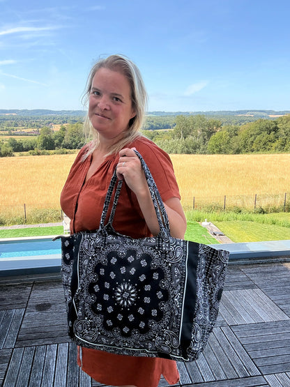 Homme/ femme portant un grand sac polochon noir à motif bandana Paisley, en extérieur près d’une terrasse et d’un paysage champêtre. Nom du produit: Sac Polochon Épaule Coton Paisley Noir – Grand Format Zippé.