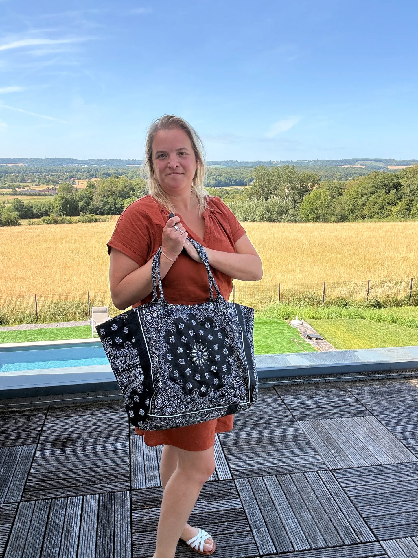 Sac polochon épaule coton noir et blanc porté par une femme debout sur une terrasse, paysage rural en arrière-plan.
