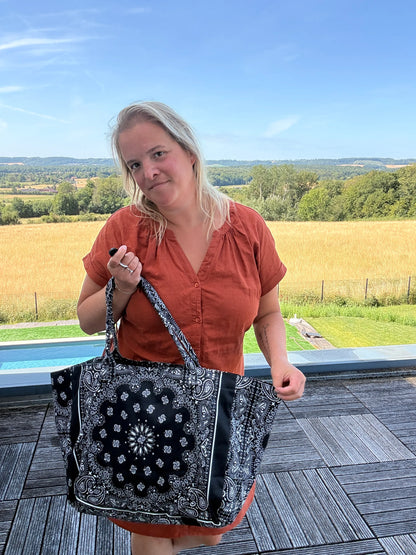 Homme/femme tenant un grand sac polochon noir à motif bandana Paisley, en extérieur sur une terrasse près d’un paysage field. Sac épaule coton paisley noir, format grand, zippé.