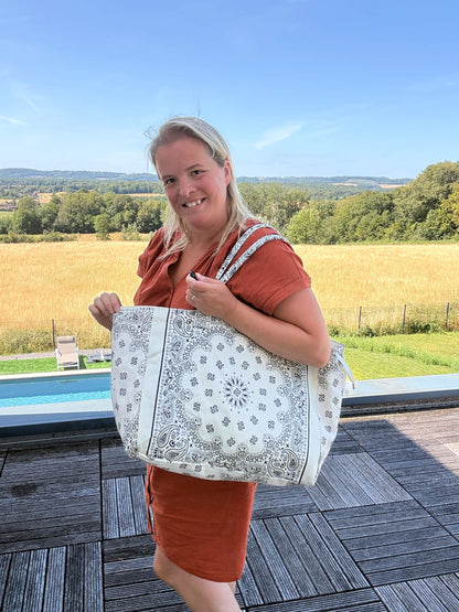 Grand sac polochon paisley blanc porté sur l’épaule par une femme, paysage champêtre en arrière-plan, sac bohème chic pour toutes les saisons.