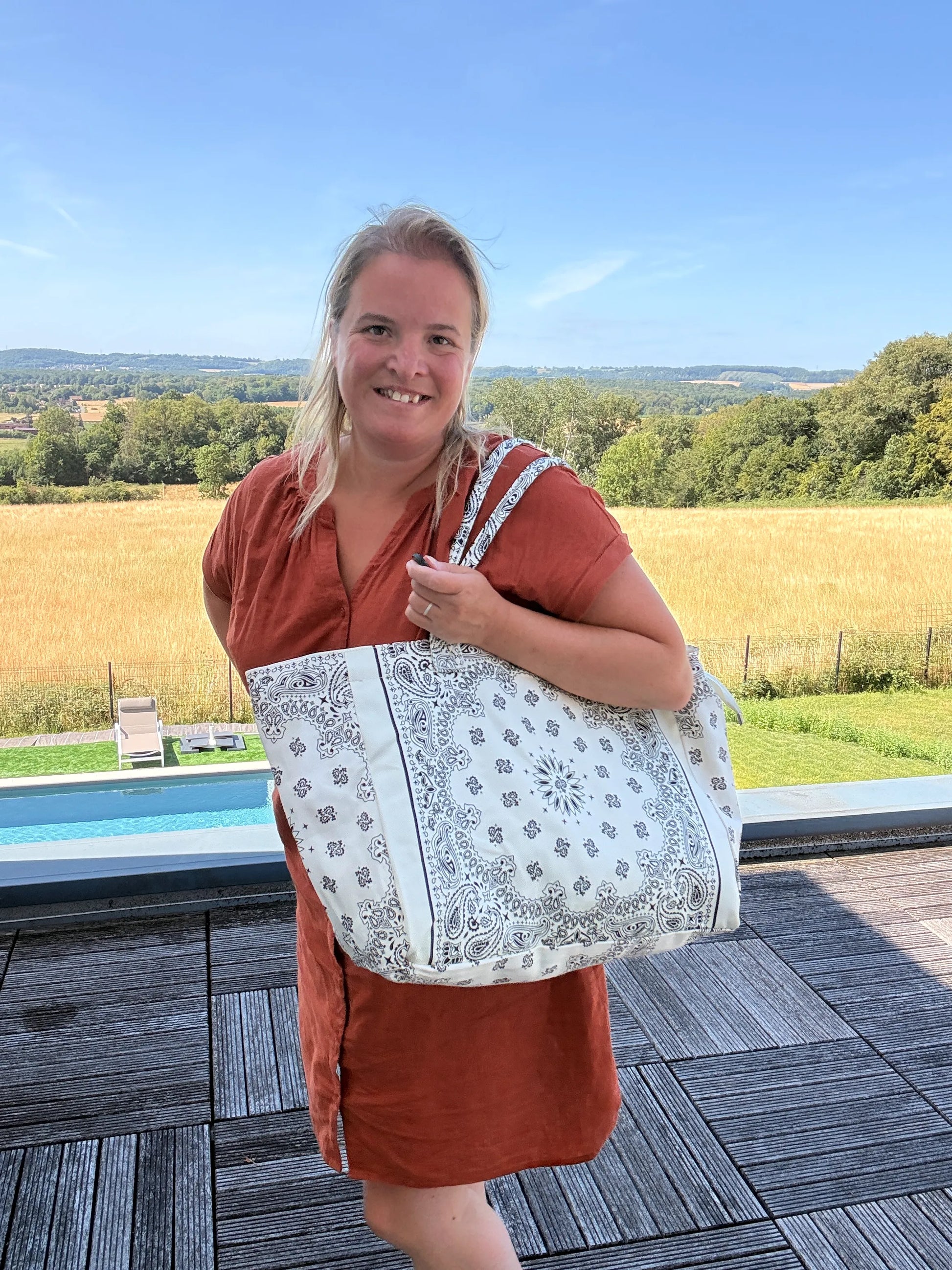Grand sac polochon Paisley blanc porté sur l’épaule par une femme en robe orange, devant une terrasse avec vue champêtre et piscine, reflet bohème chic.