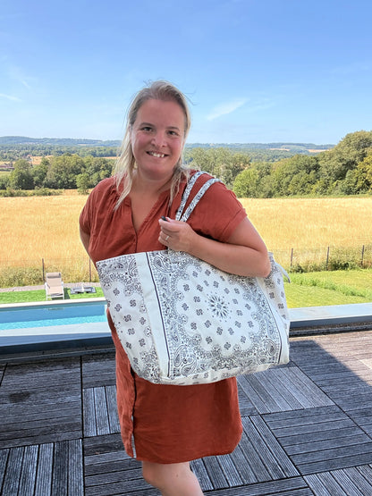 Grand sac polochon Paisley blanc porté sur l’épaule par une femme en robe orange, devant une terrasse avec vue champêtre et piscine, reflet bohème chic.