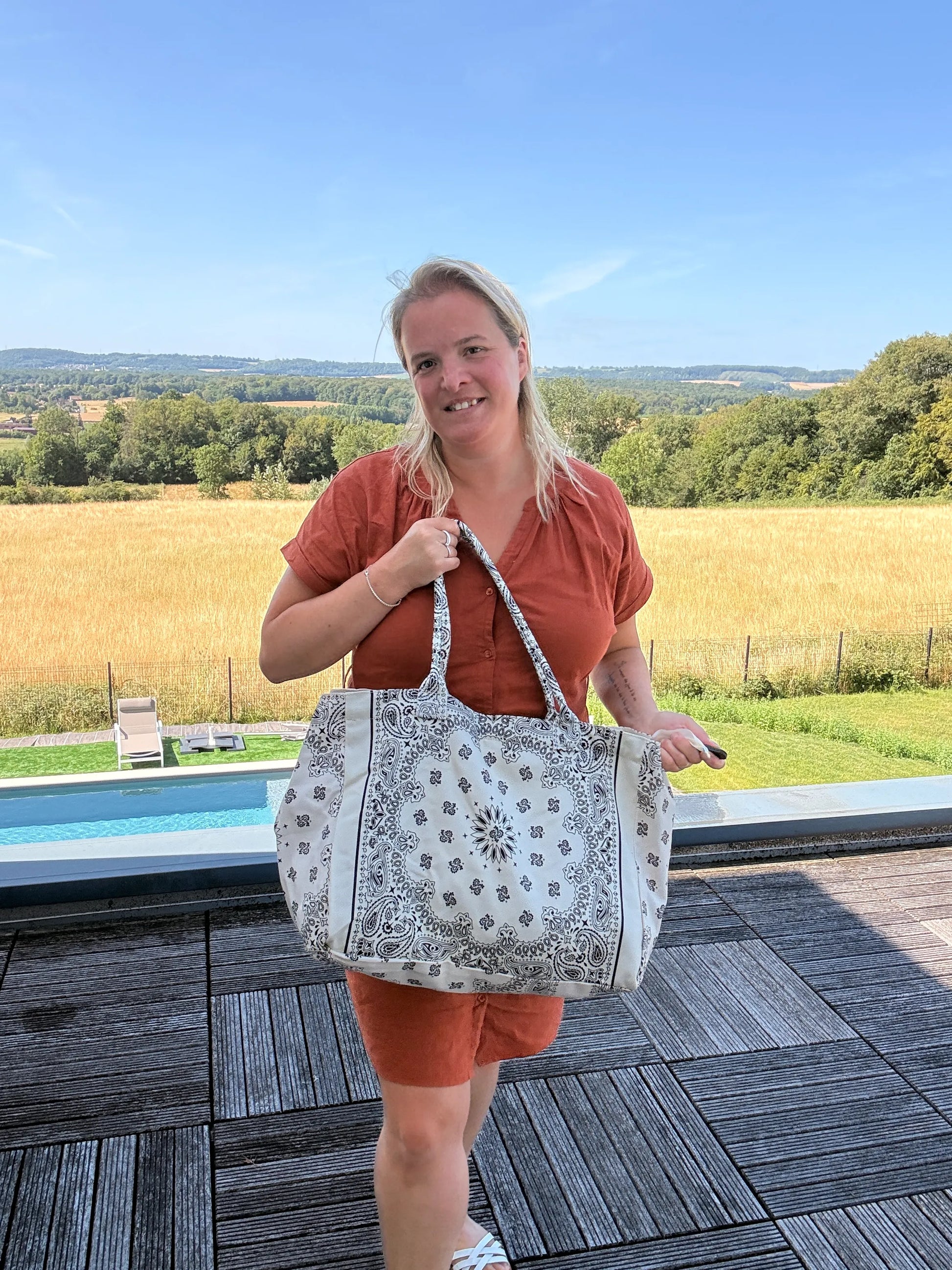Grande sac polochon paisley blanc porté par une femme en tenu orange, sur fond de paysage rural et piscine.