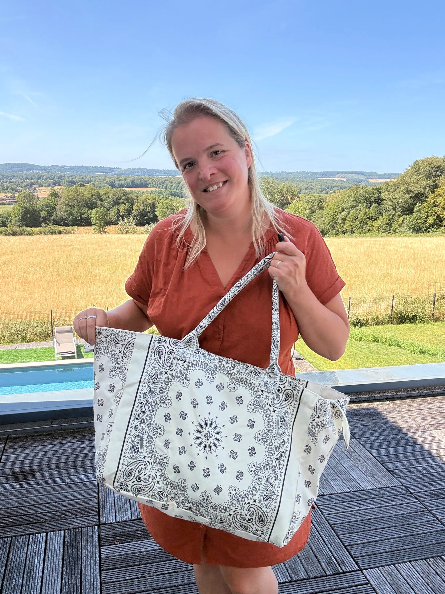 Grand Sac Polochon Paisley Blanc, femme souriante tenant un grand sac bohème chic à motifs paisley blanc, devant paysage champêtre.