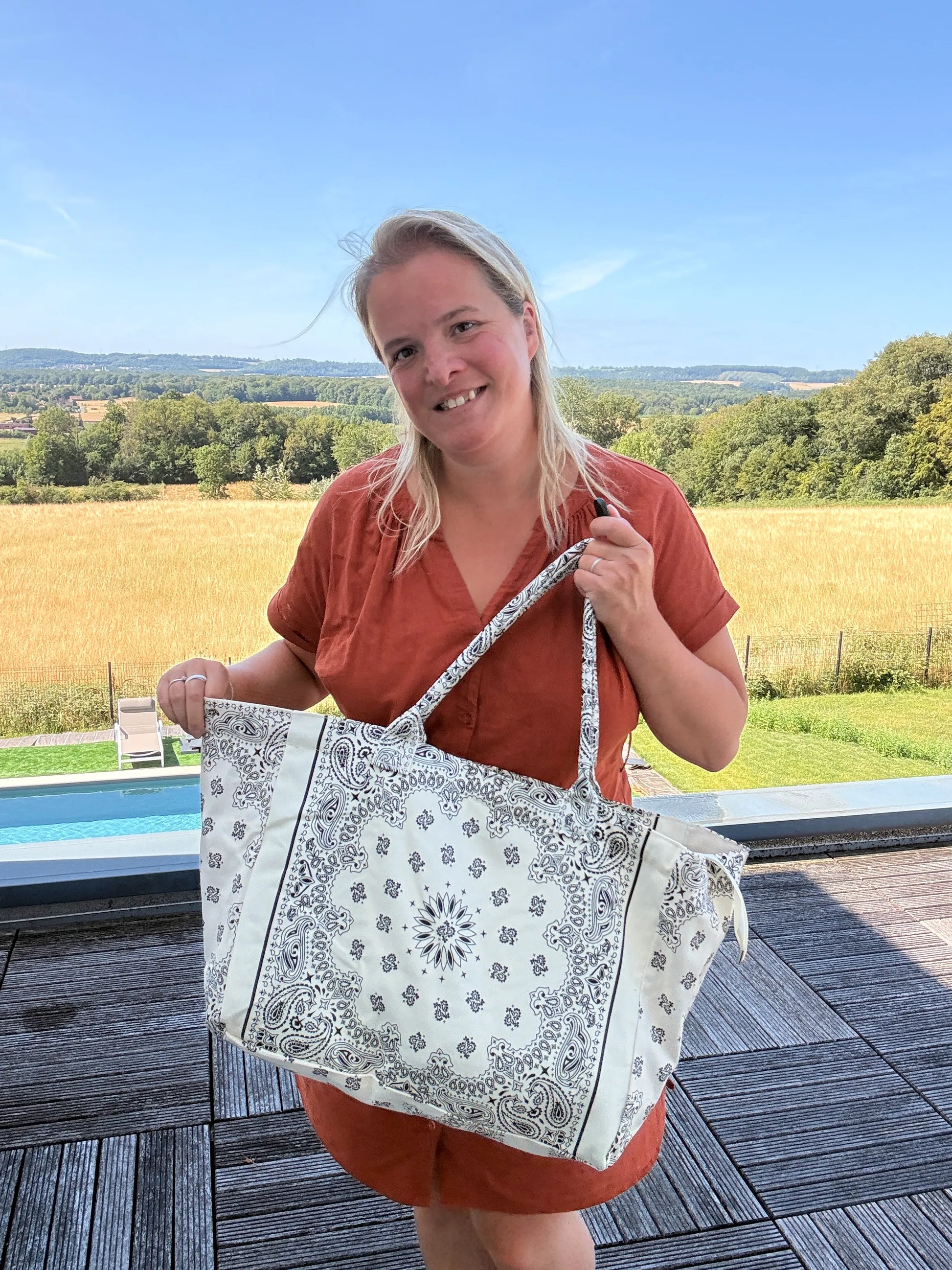 Grand Sac Polochon Paisley Blanc, femme souriante tenant un grand sac bohème chic à motifs paisley blanc, devant paysage champêtre.