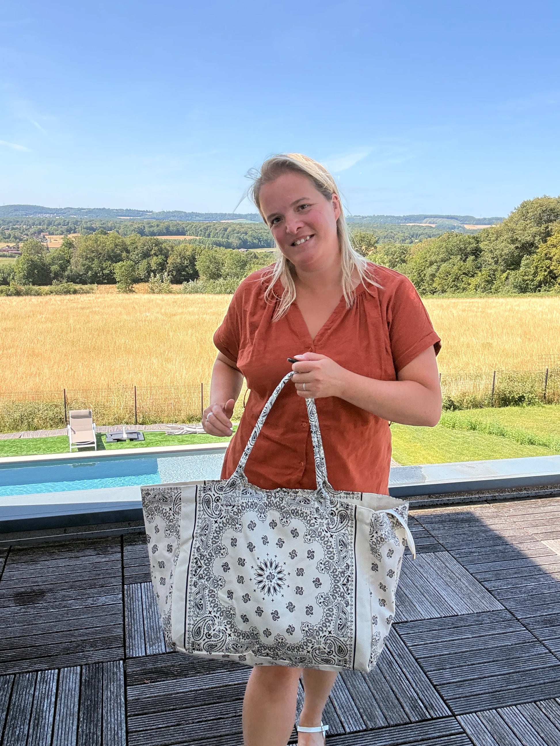 Homme ou femme tenant un grand sac polochon blanc à motif paisley, posé en main sur une plage arrière avec paysage rural en arrière-plan. Grand Sac Polochon Paisley Blanc, Le Sac Bohème Chic de Toutes les Saisons.