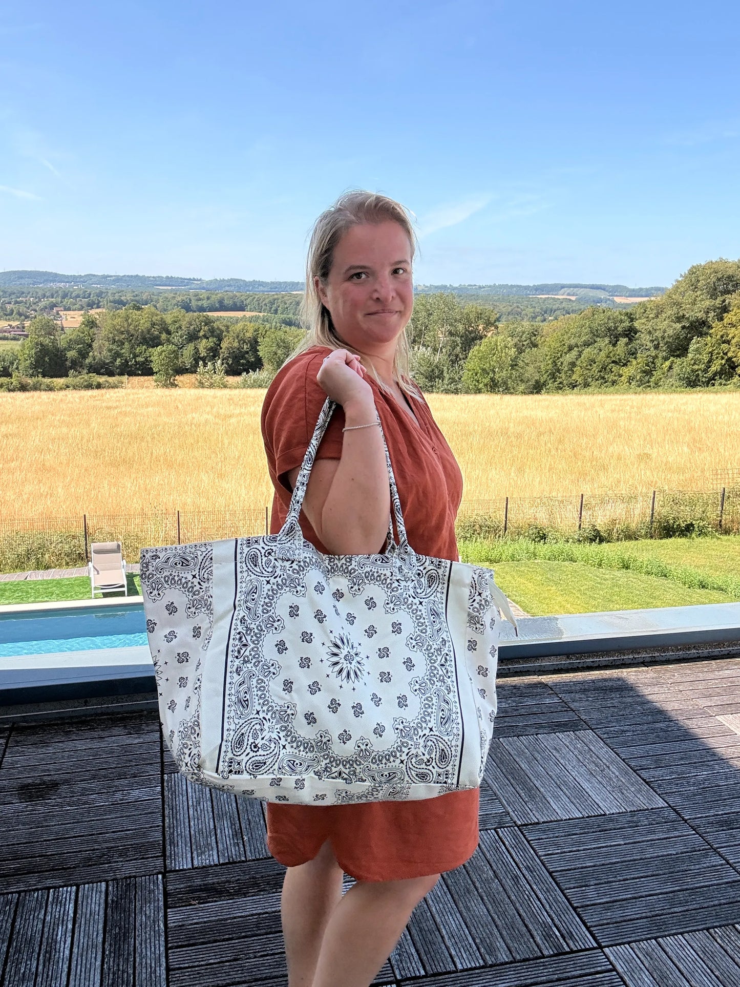 Grand sac polochon blanc paisley porté par une femme en robe orange, paysage champêtre en fond, près d’une piscine.