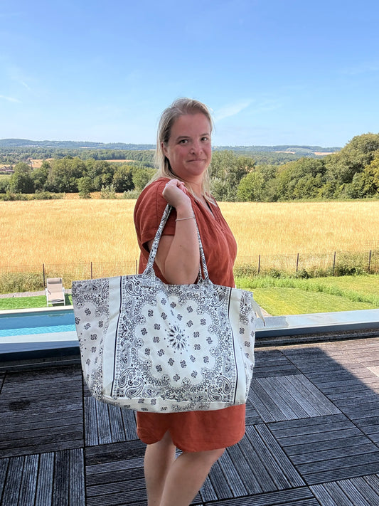 Grand sac polochon blanc paisley porté par une femme en robe orange, paysage champêtre en fond, près d’une piscine.