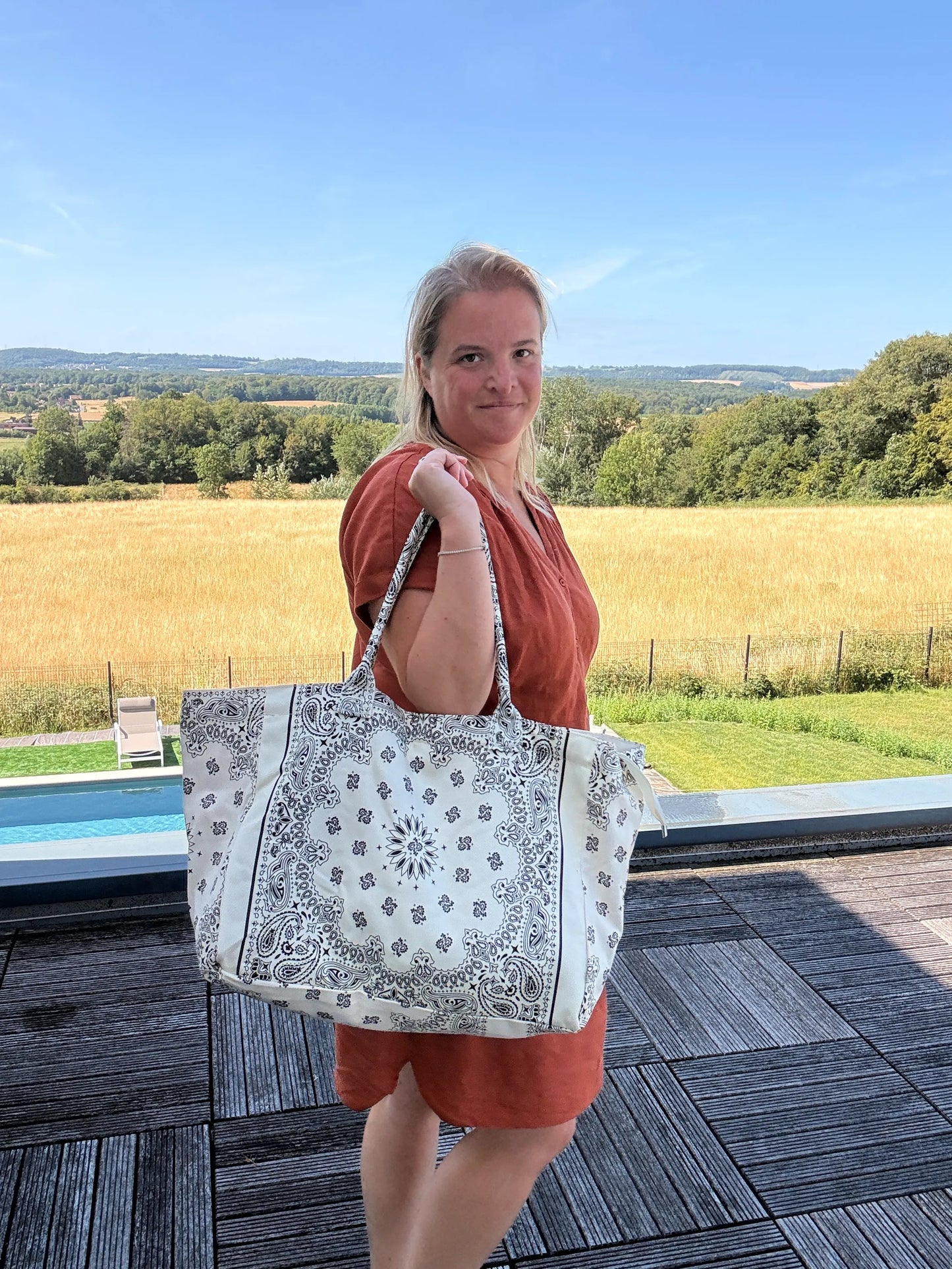 Grand sac polochon paisley blanc porté par une femme en robe orange, paysage champêtre en arrière-plan et piscine à proximité.