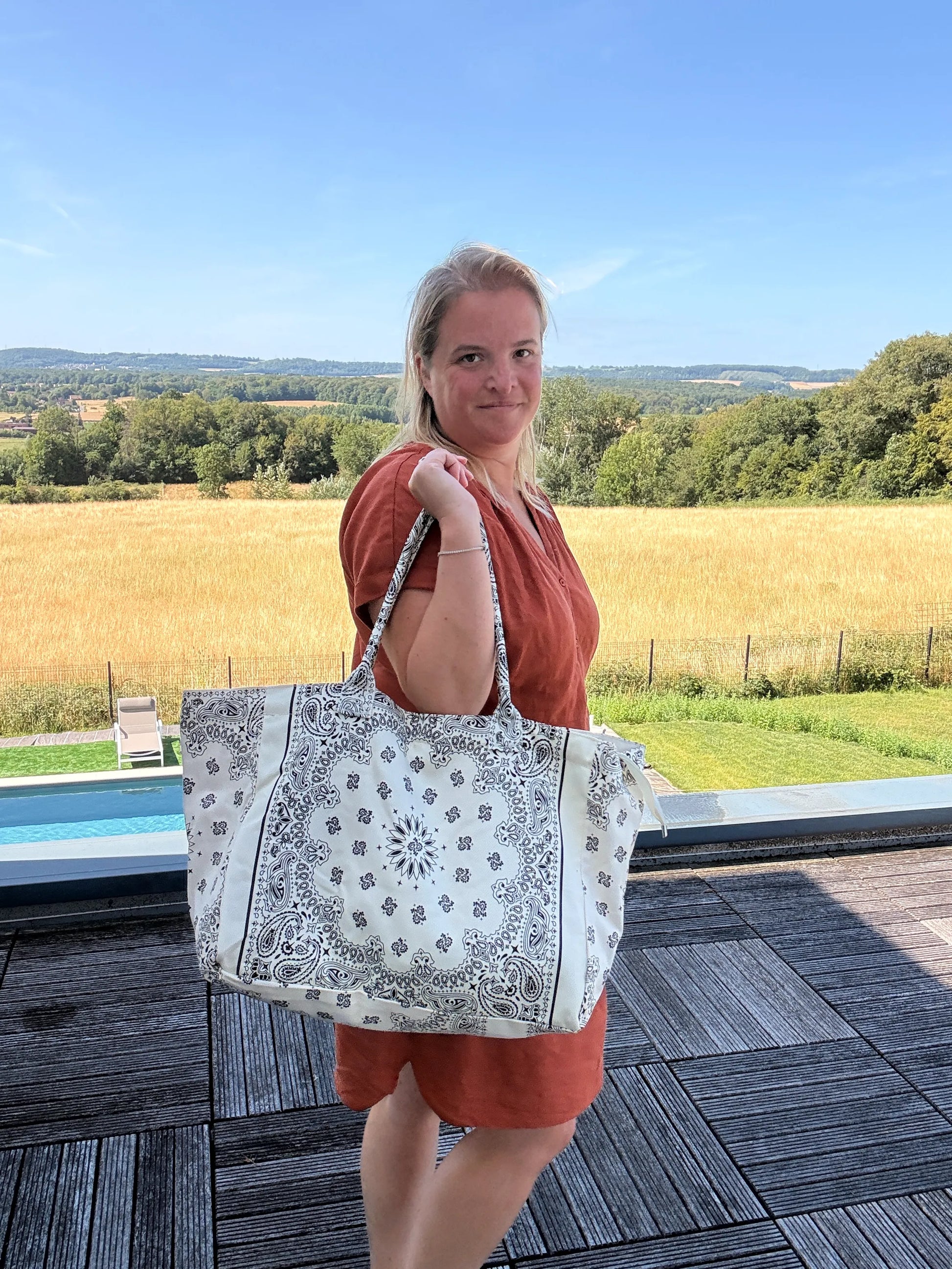Grand sac polochon paisley blanc porté par une femme en robe orange, paysage champêtre en arrière-plan et piscine à proximité.