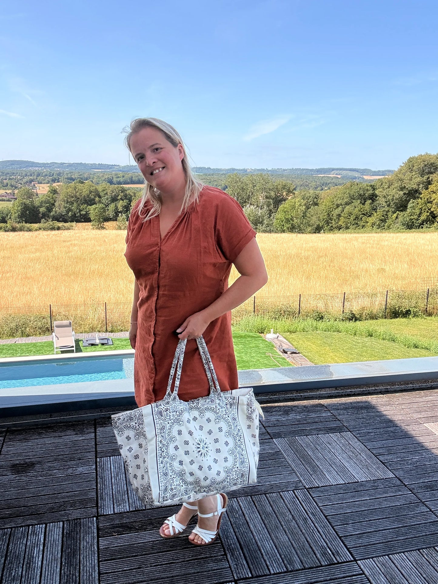 Homme ou femme souriante tenant un grand sac polochon blanc à motif paisley, en extérieur sur terrasse avec paysage rural derrière.