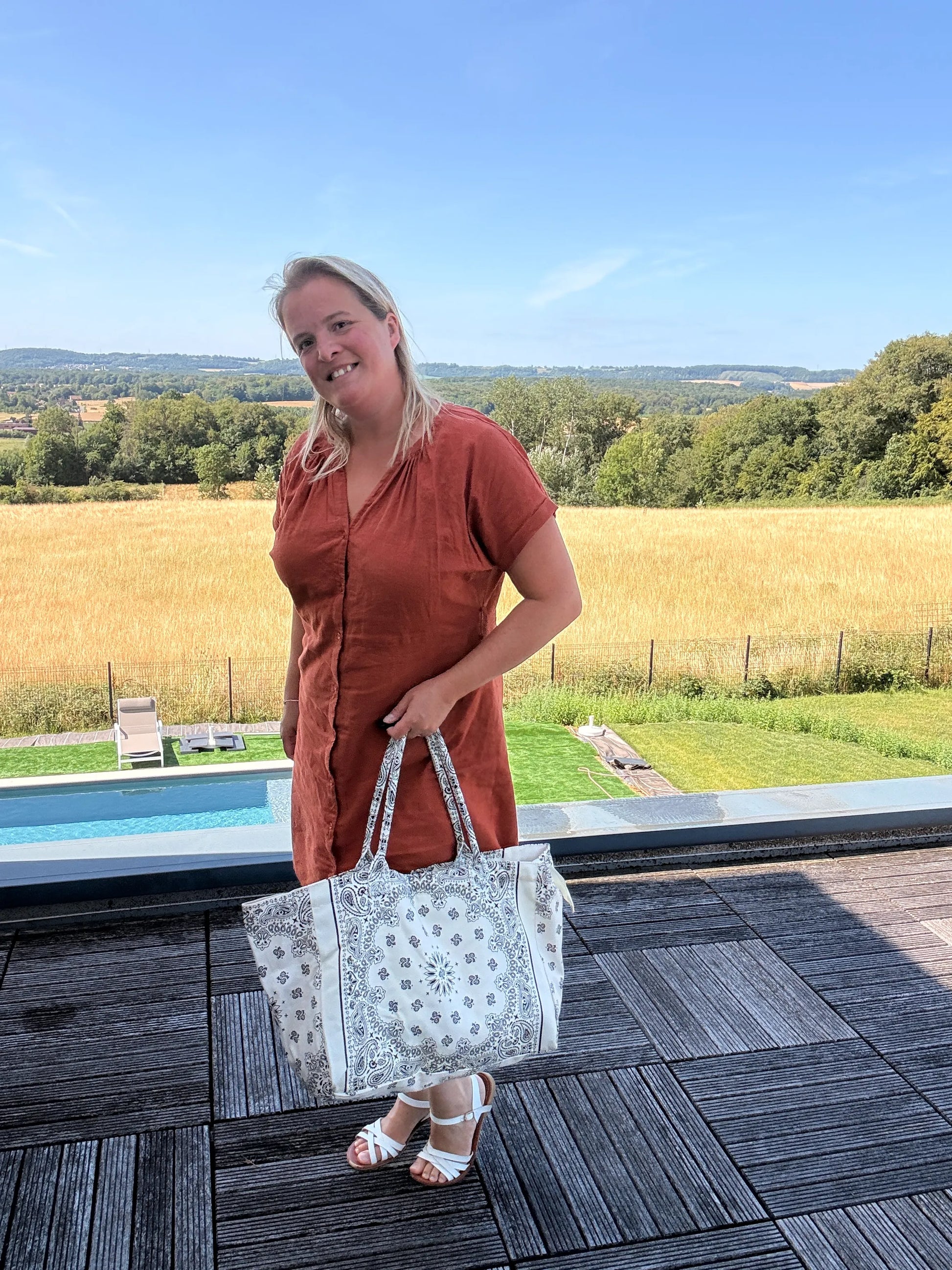 Homme ou femme souriante tenant un grand sac polochon blanc à motif paisley, en extérieur sur terrasse avec paysage rural derrière.