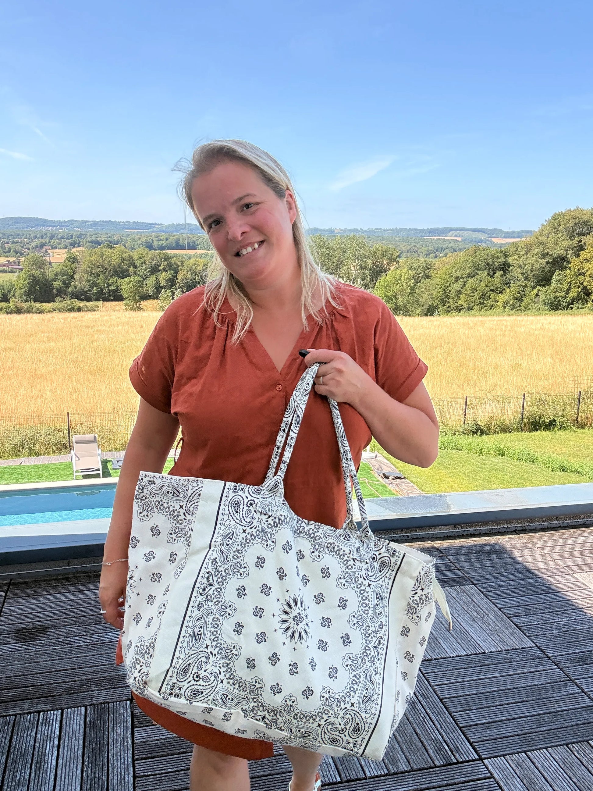 Grand sac polochon blanc à imprimé paisley porté par une femme en robe orange devant un paysage champêtre et piscine.