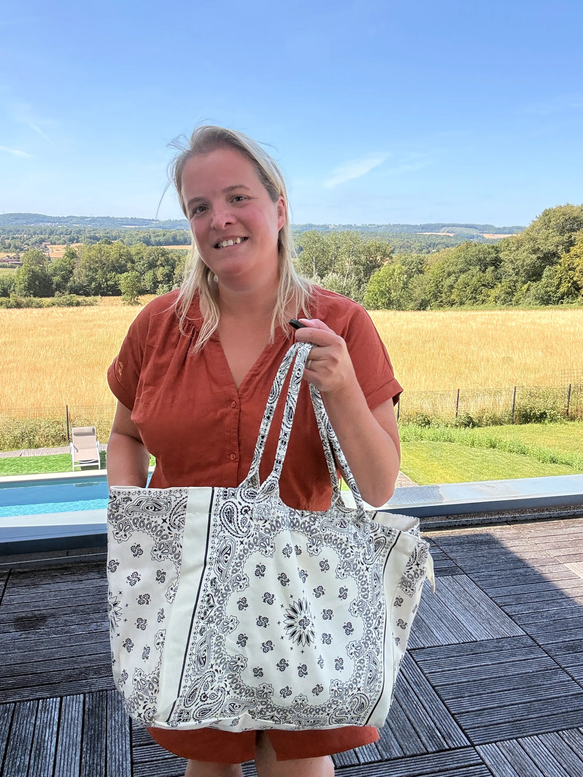 Homme ou femme tenant un grand sac polochon blanc à motifs Paisley, tenu à la main près d’une terrasse, paysage rural en arrière-plan. Grand sac bohème chic pour toutes les saisons.