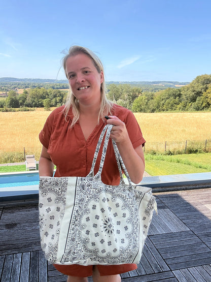 Homme ou femme tenant un grand sac polochon blanc à motifs Paisley, tenu à la main près d’une terrasse, paysage rural en arrière-plan. Grand sac bohème chic pour toutes les saisons.