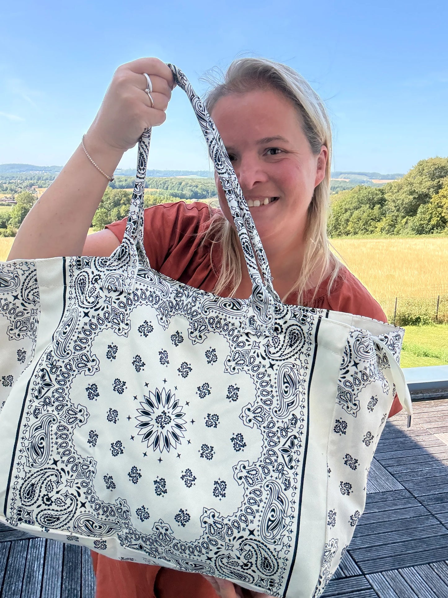 Grand Sac Polochon Paisley Blanc, femme souriante tenant un grand sac bandoulière imprimé paisley blanc et noir, paysage champêtre en arrière-plan.