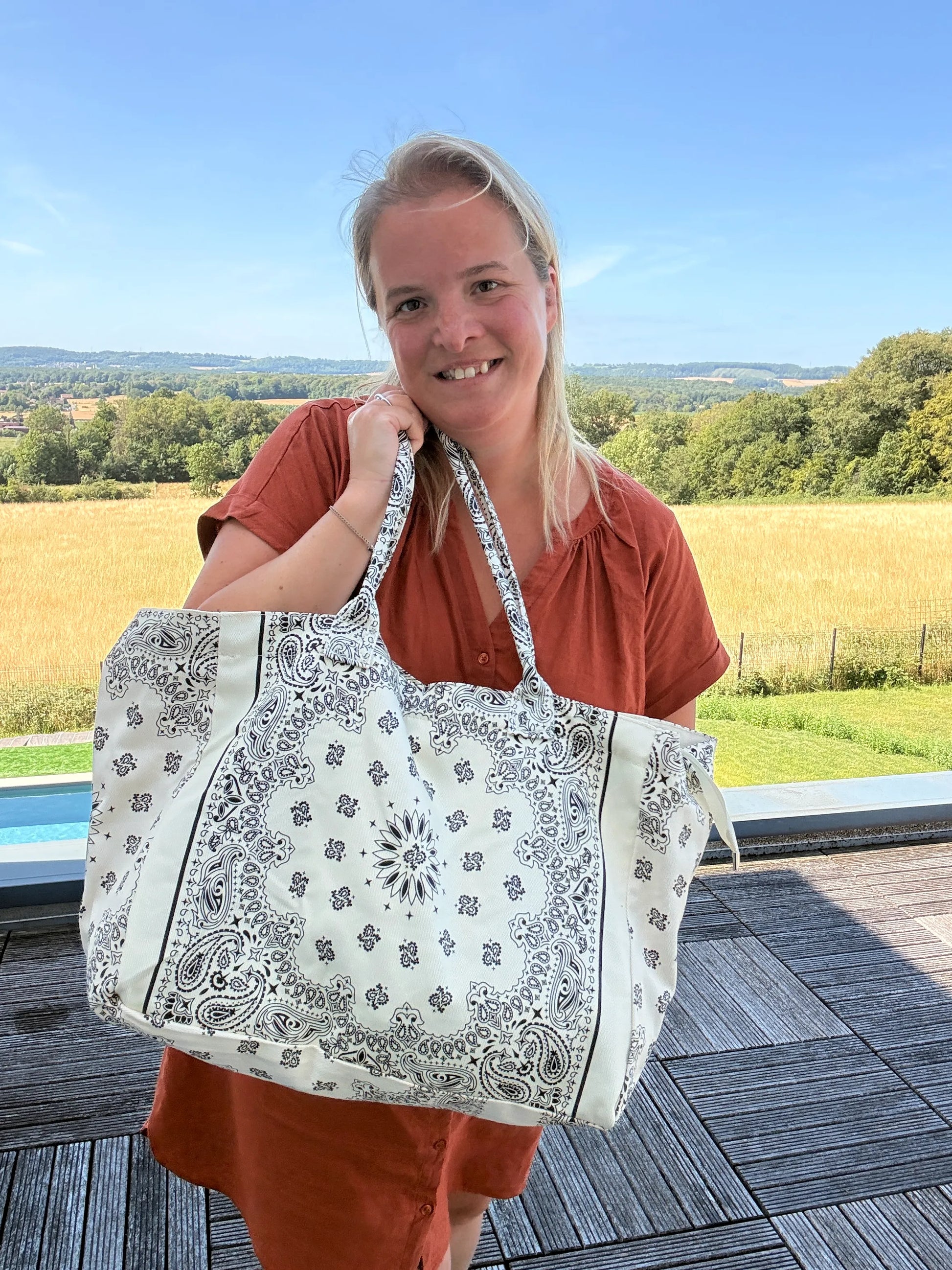 Grande sac polochon blanc à motif bandana noir porté par une femme en robe rouge, sourire, paysage champêtre en arrière-plan.