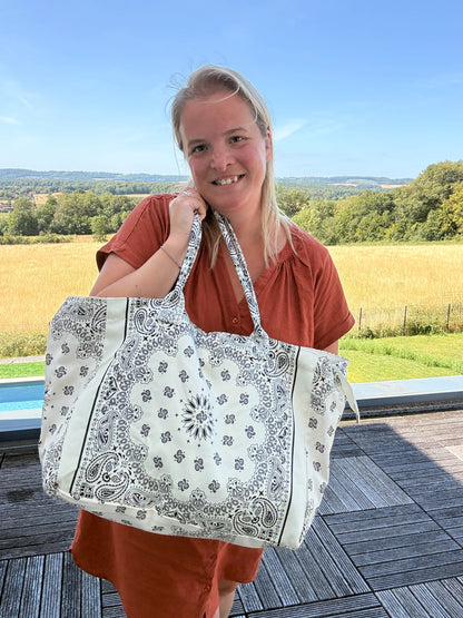 Grande sac polochon blanc à motif bandana noir porté par une femme en robe rouge, sourire, paysage champêtre en arrière-plan.