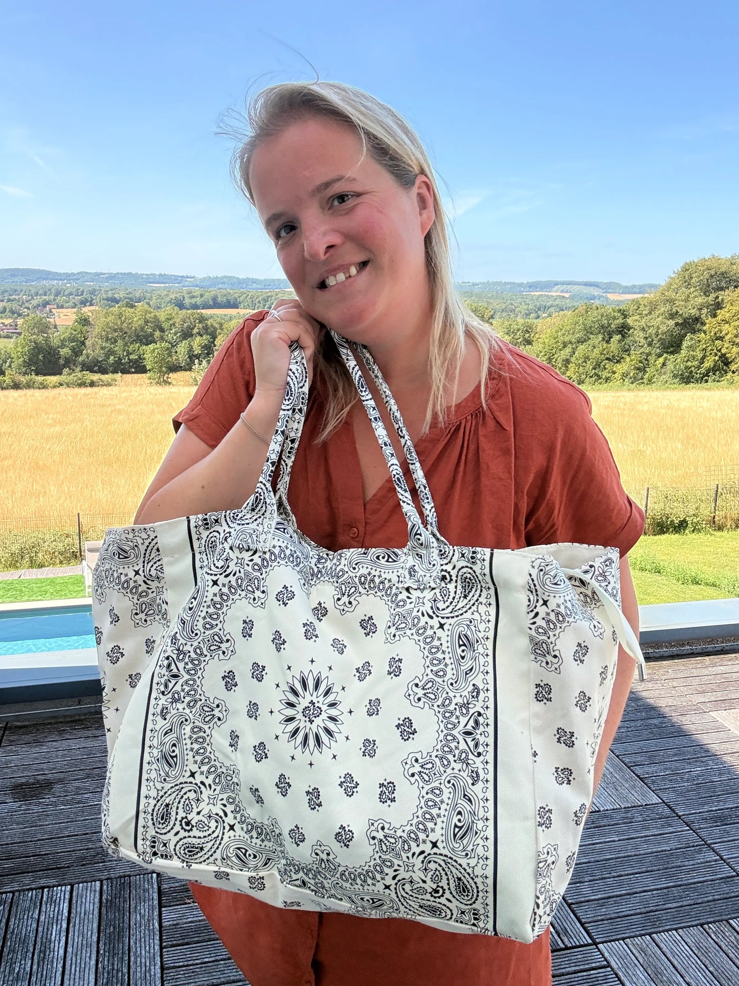 Sophie souriante tenant un grand sac polochon blanc et noir à motif bandana paisley, en extérieur près d’un paysage champêtre.